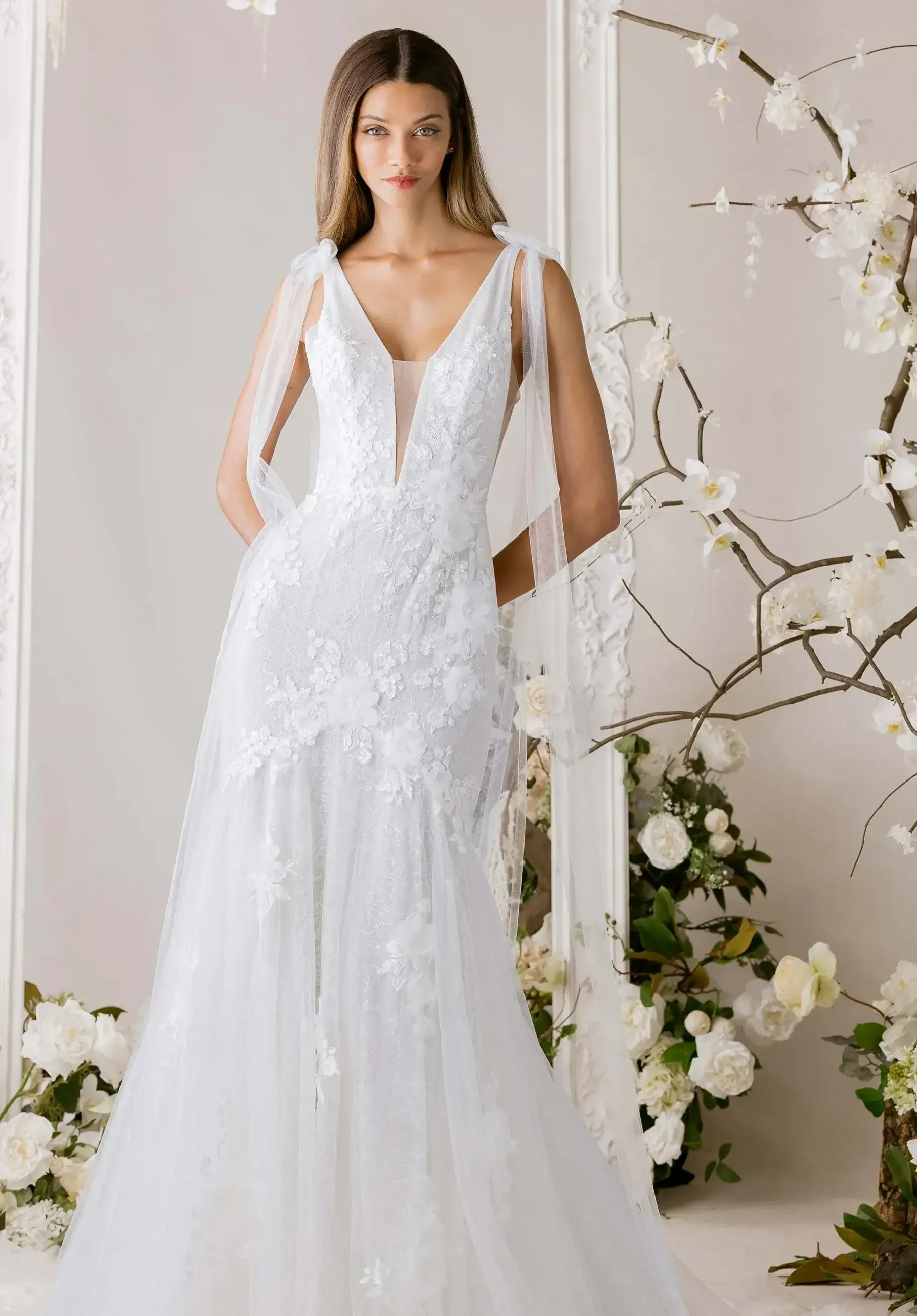 Woman in elegant white lace wedding gown with deep V-neckline and sheer shoulder accents stands in floral setting, exuding a romantic, serene vibe.