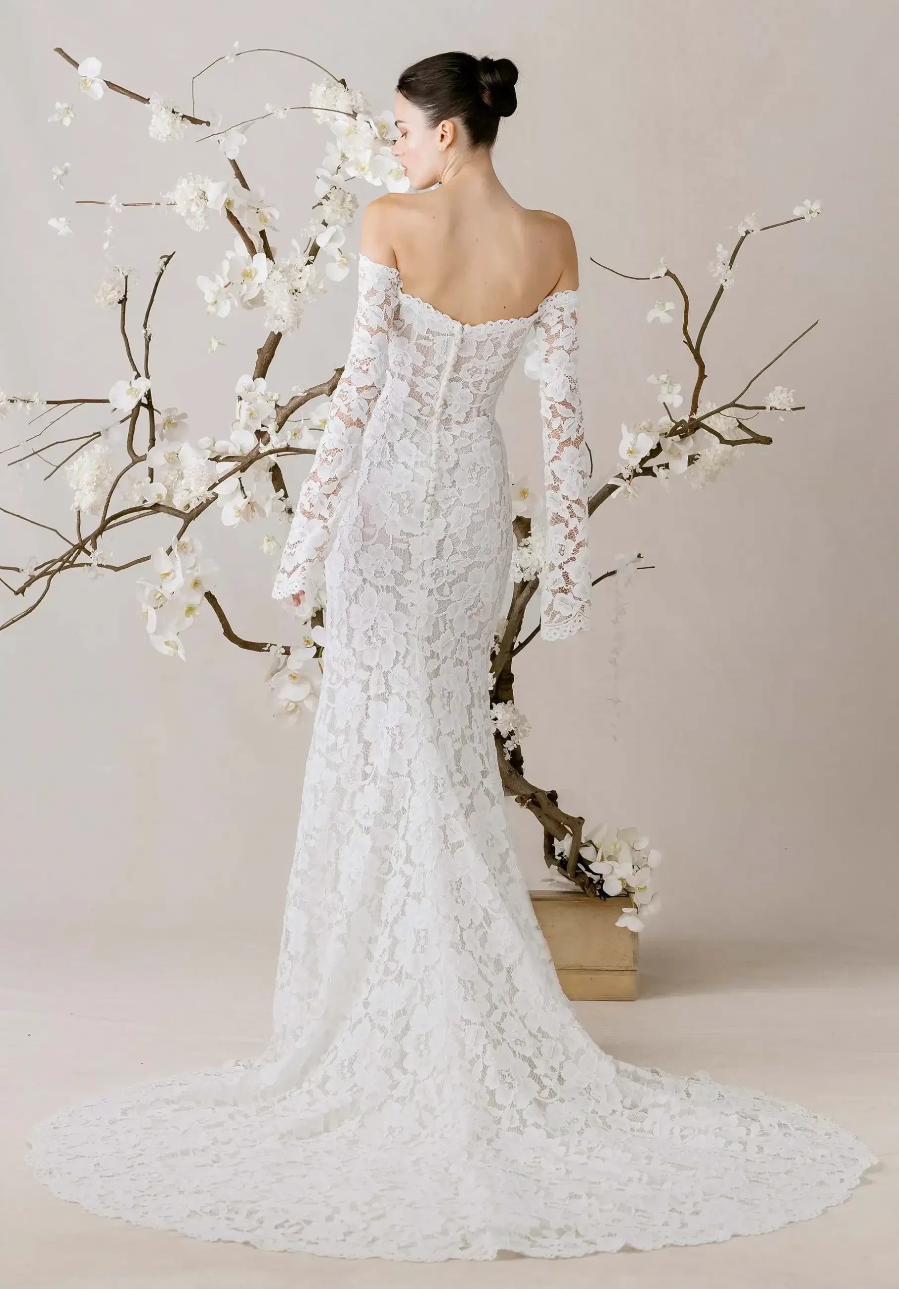 A woman in a white lace off-the-shoulder gown stands with her back turned, near a decorative branch with white flowers, creating an elegant, serene scene.
