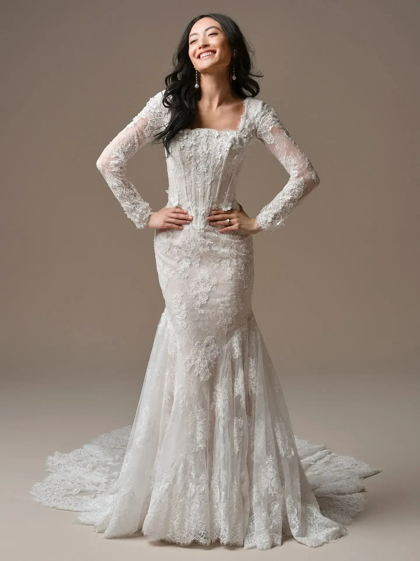 Smiling woman in a lace wedding dress with long sleeves and a mermaid cut stands confidently against a neutral background, exuding joy and elegance.