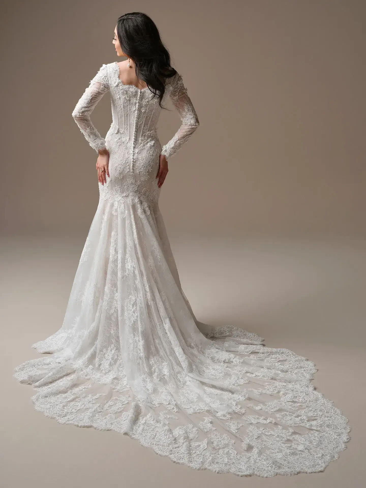 A woman stands with her back to the camera, wearing a fitted white lace wedding dress with long sleeves and a flowing train, exuding elegance and romance.