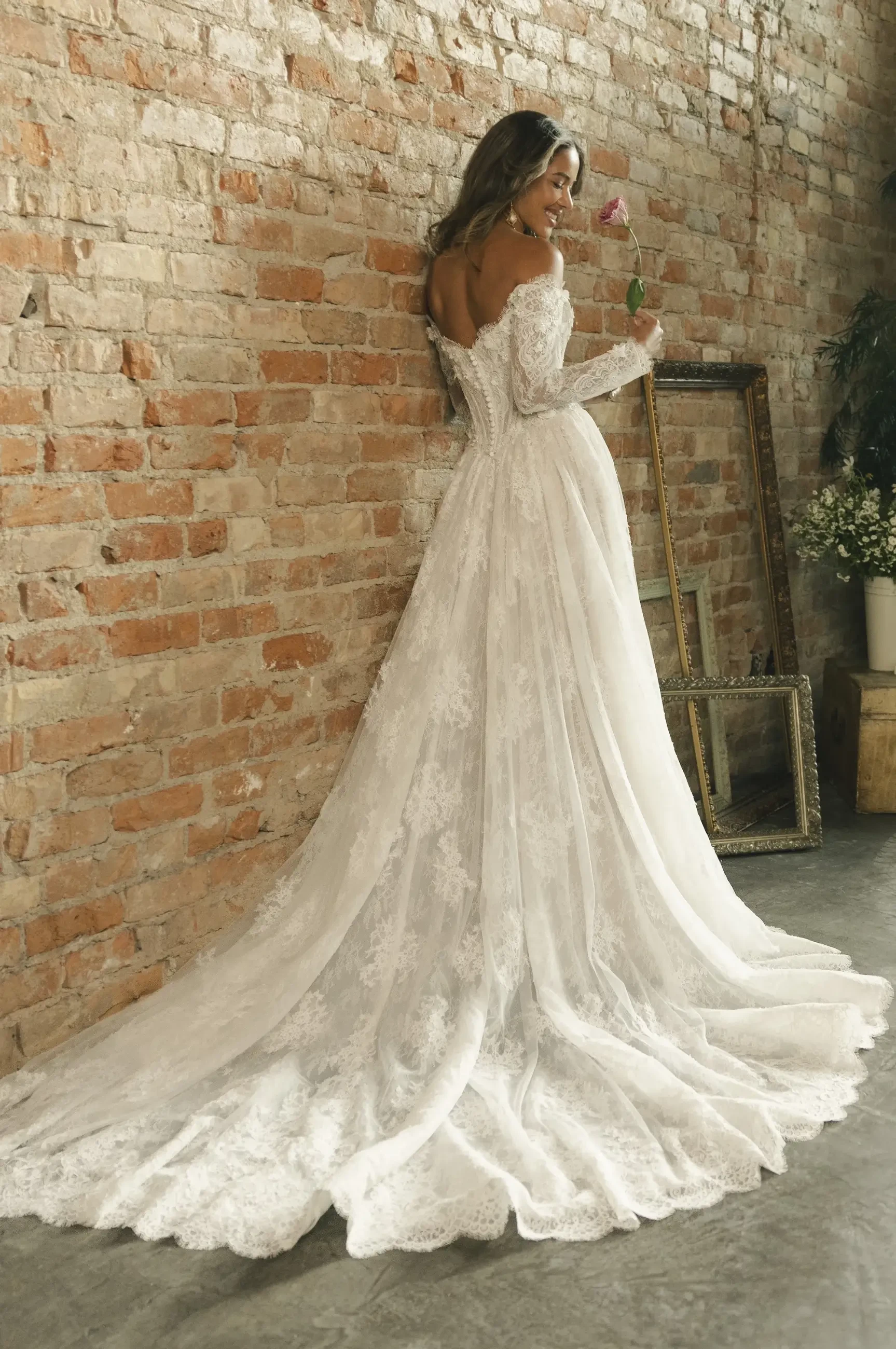 Bride in an elegant lace wedding gown stands by a rustic brick wall, holding a pink rose. The dress features a long train and off-the-shoulder sleeves.