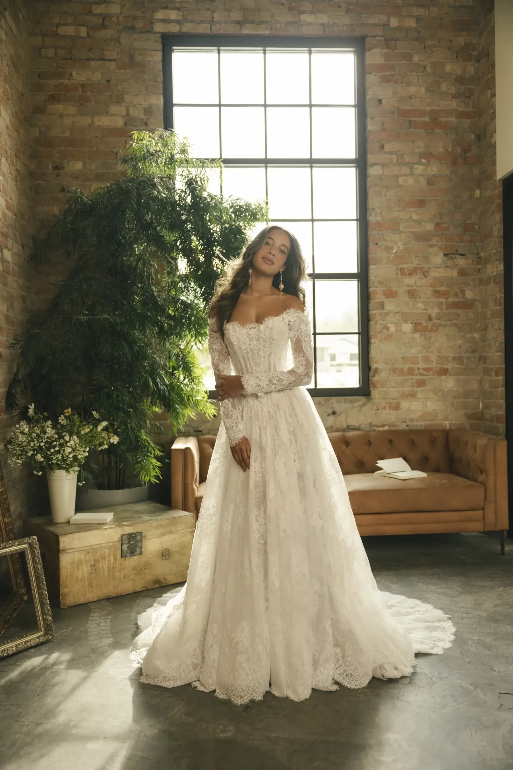 A bride in a lace wedding gown stands in a rustic room with brick walls, a large window, and a potted plant. The atmosphere is serene and elegant.