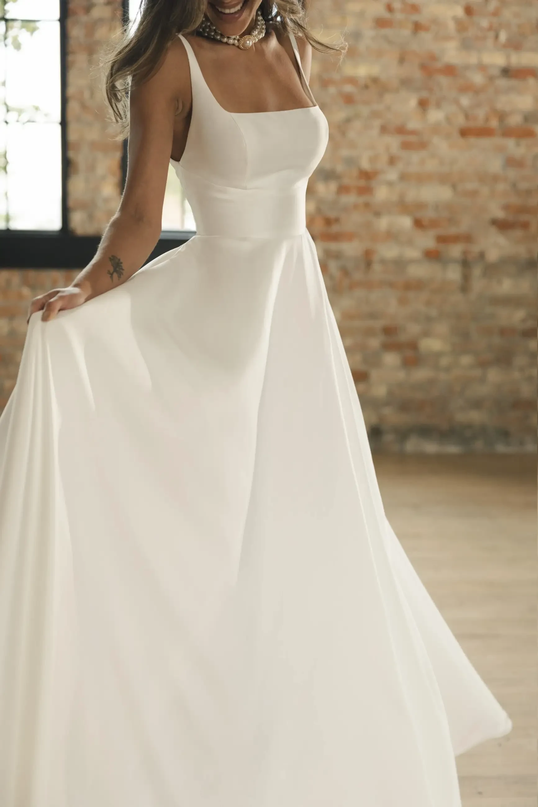 A woman in a flowing white wedding dress stands in a rustic room with exposed brick walls and large windows, exuding elegance and joy.