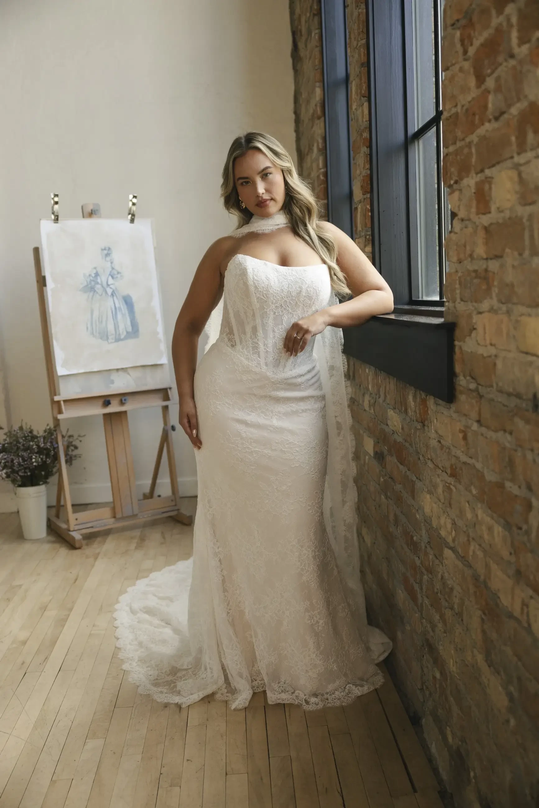 A woman in an elegant strapless wedding dress leans against a brick wall by a large window. An art easel with a blue sketch and a potted plant are in the background, creating a serene and sophisticated atmosphere.