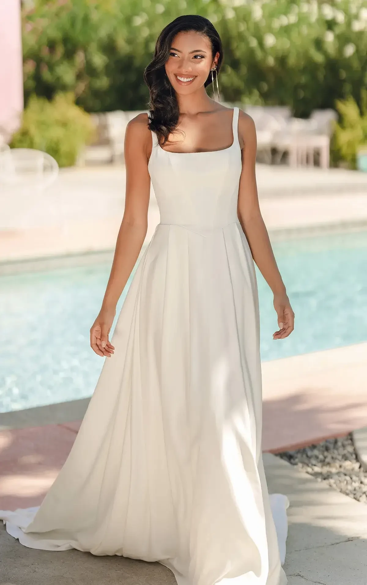 A woman in a flowing white gown stands elegantly by a pool, surrounded by lush greenery. She is smiling softly, conveying a serene, joyful mood.
