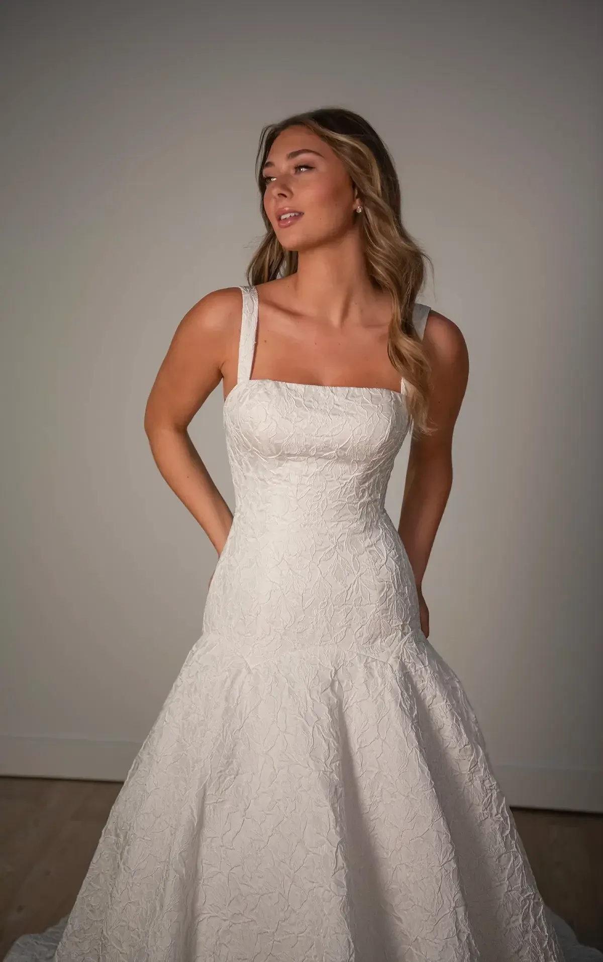 A woman in an elegant white textured gown stands confidently against a neutral background. Her expression is serene, and she exudes grace and poise.